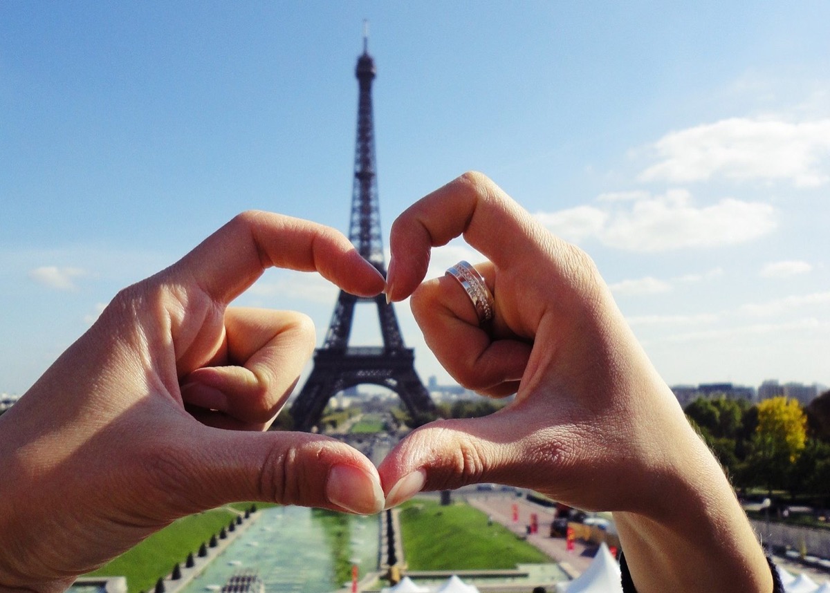 A couple creating a heart shape with their hands in front of the Eiffel Tower