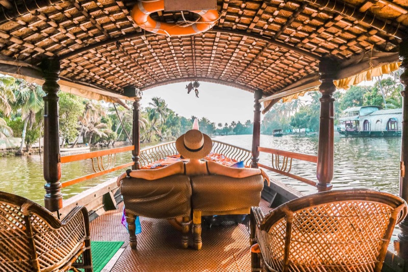 Kerala backwaters in Alleppey — a traditional wooden houseboat gliding through palm-fringed canals at golden hour, surrounded by lush tropical greenery