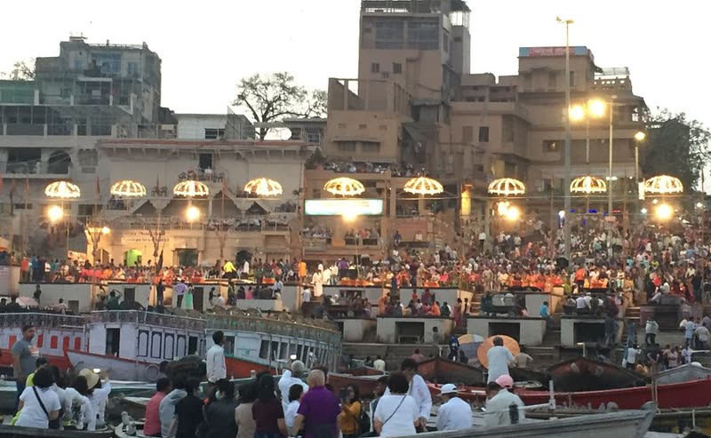Evening Ganga Aarti ceremony at Varanasi's Dashashwamedh Ghat — priests in saffron robes performing synchronized rituals with brass oil lamps while smoke and lights reflect on the Ganges river