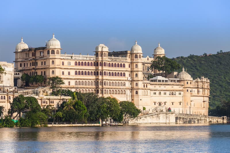 Udaipur City Palace complex — the massive lakeside fortress rising above Lake Pichola with its domes and towers reflecting on the water at golden hour
