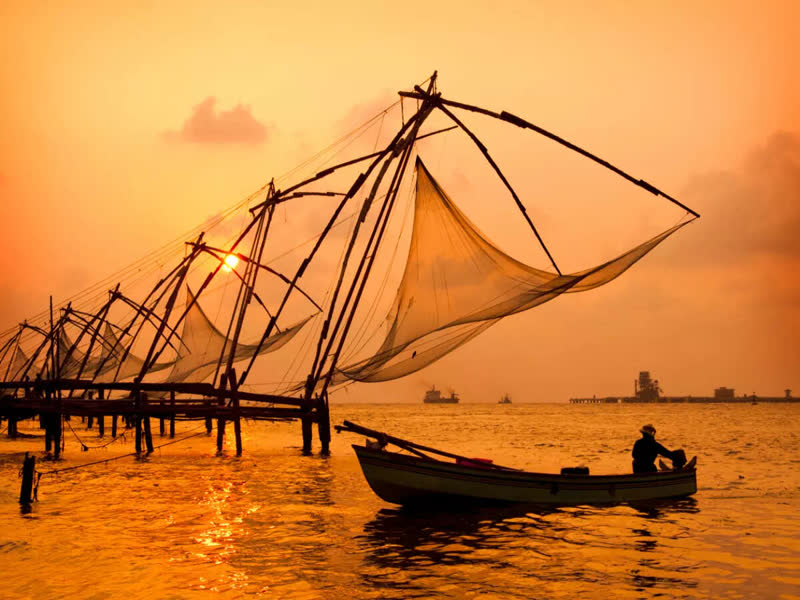 Chinese fishing nets at Fort Kochi at dusk — the iconic cantilevered fishing structures silhouetted against an orange sunset sky over the Kochi waterfront with boats in the harbor