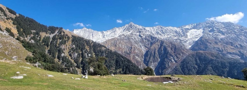 The Dhauladhar mountain range viewed from Triund ridge near Dharamsala — snow-capped peaks rising dramatically above the valley, with trekkers at the campsite looking up at the Himalayan wall