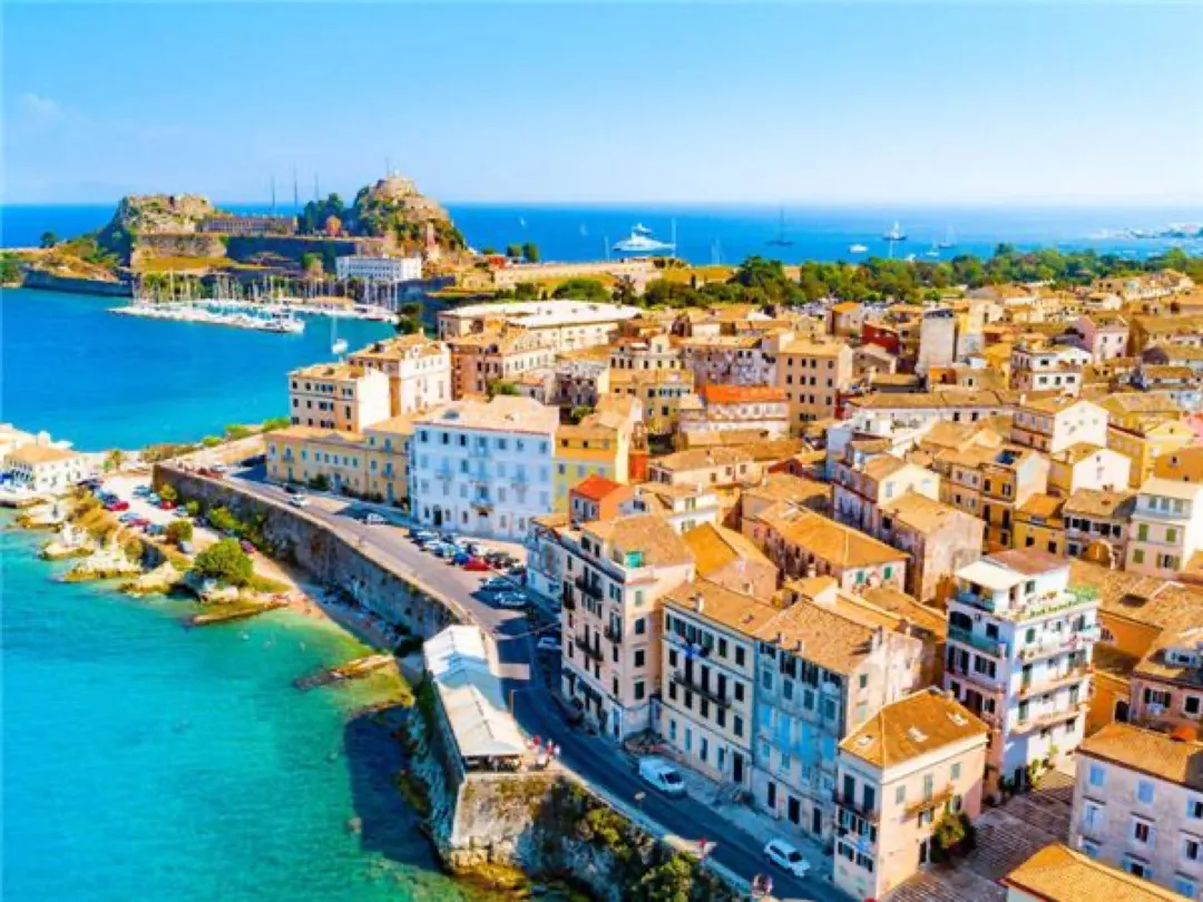 Venetian-era pastel buildings of Corfu Old Town line the vibrant Spianada square under a clear blue sky, bustling with locals and visitors.
