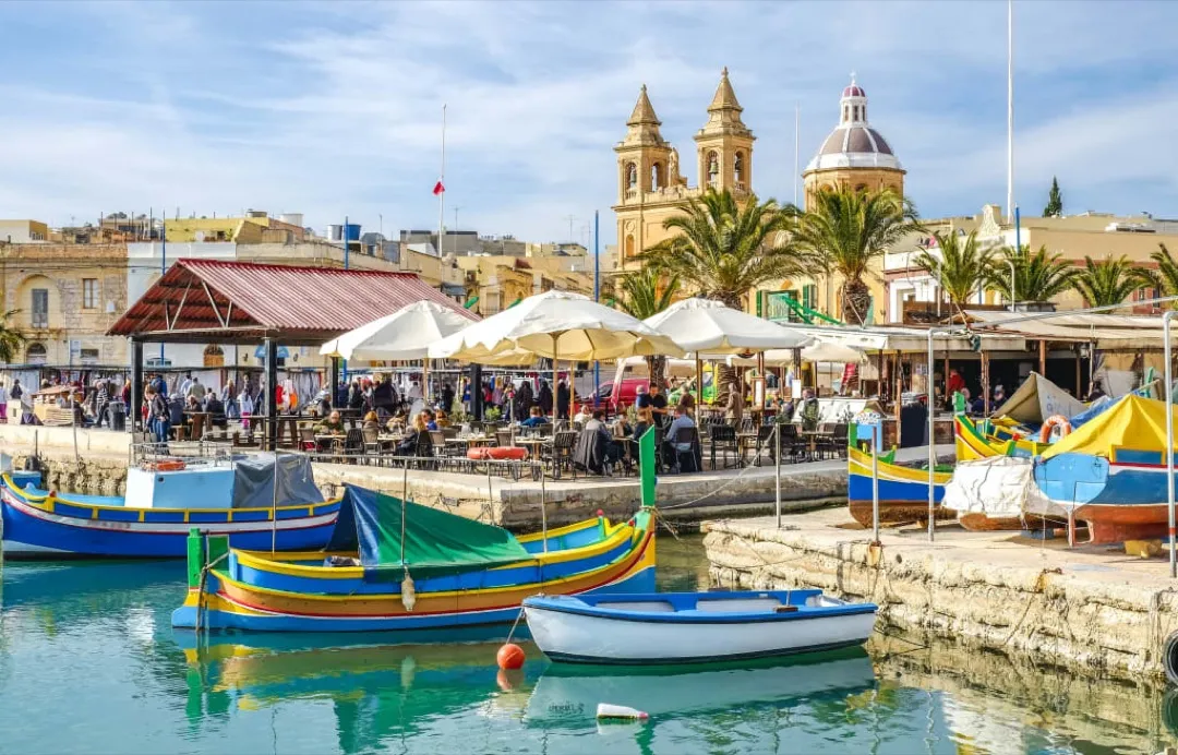 A traditional Maltese luzzu fishing boat gently bobs in Marsaxlokk Bay, framed by pastel-colored houses.