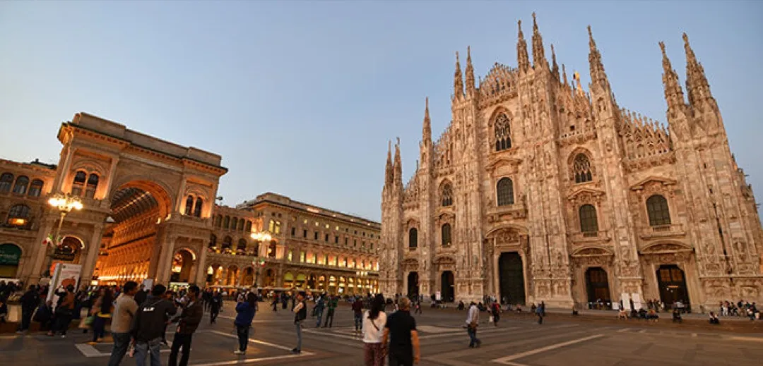 The intricate Gothic facade of Milan Cathedral (Duomo di Milano) rises majestically against a bright sky, surrounded by bustling Piazza del Duomo.