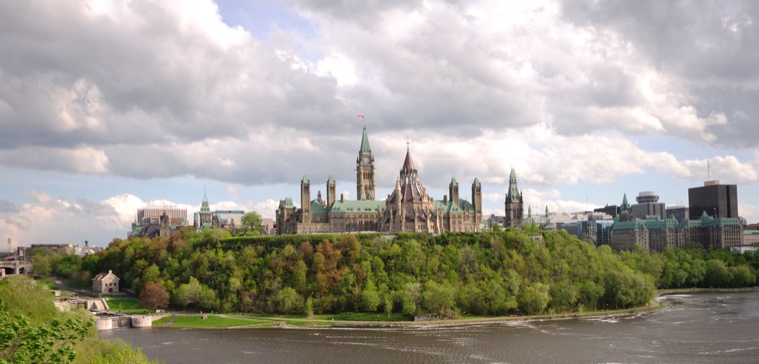 Parliament Hill's grand Gothic revival buildings stand proudly against a clear blue sky, overlooking the serene Ottawa River.