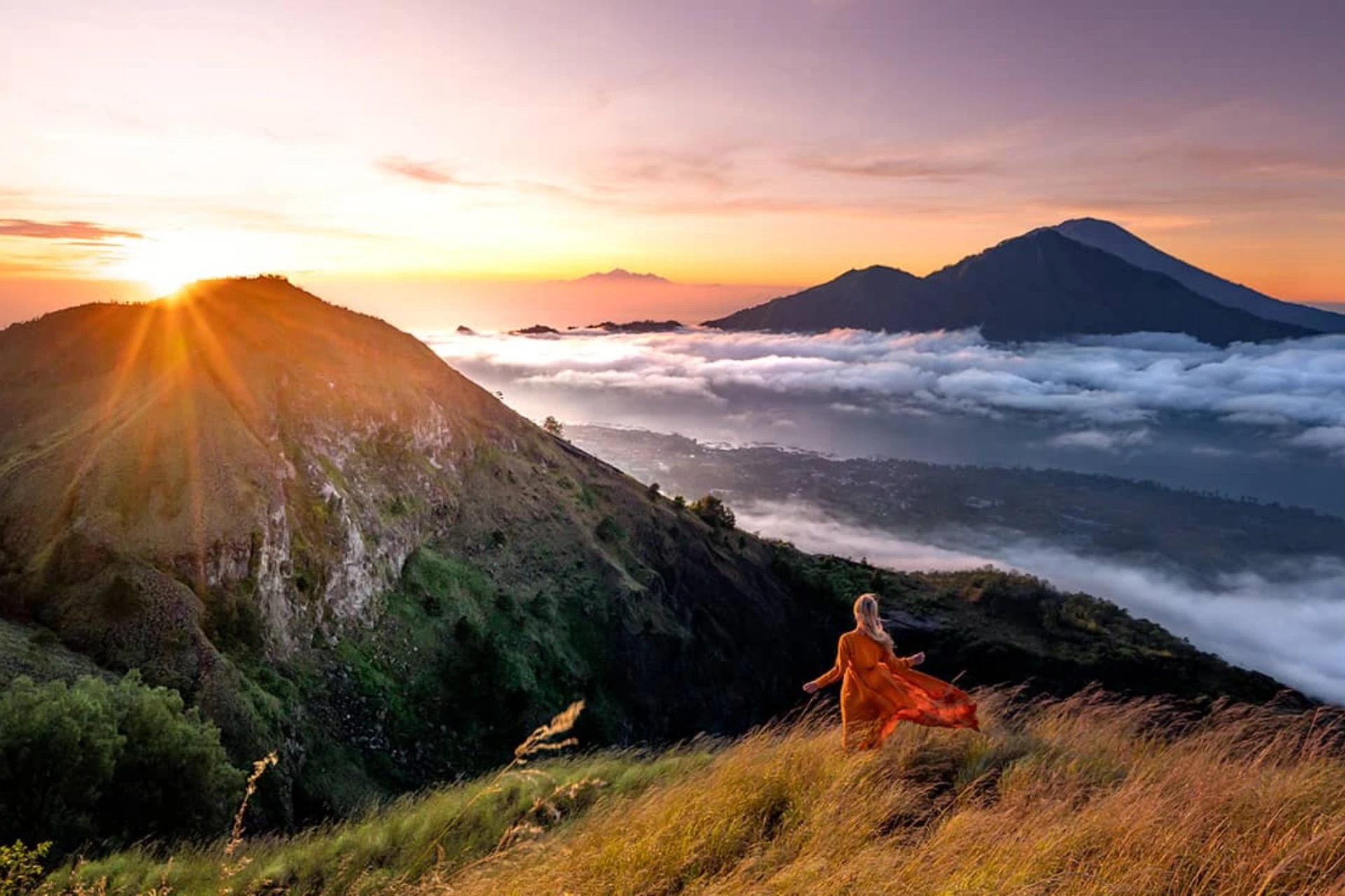 Hiker silhouetted against dramatic Mount Batur volcano sunrise with sunburst and volcanic landscape, Bali